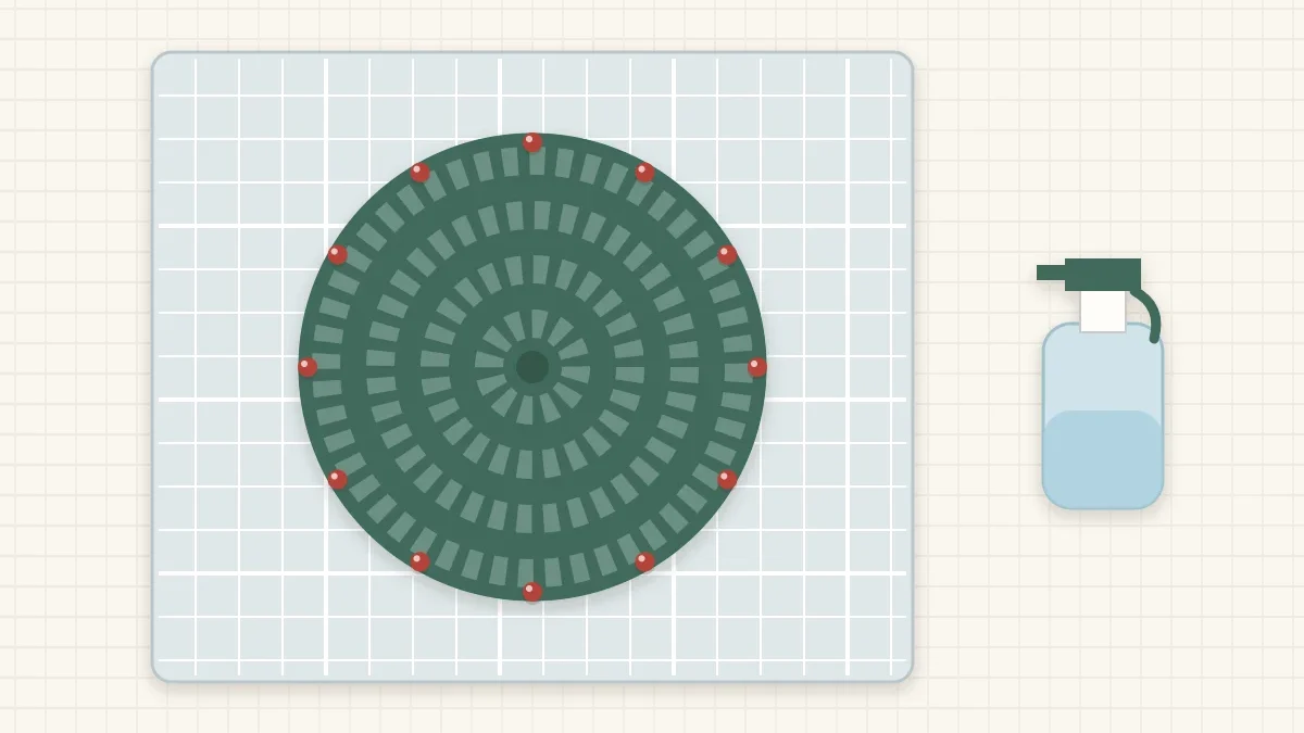 A round coaster pinned to a gridded blocking mat with a spray bottle beside it
